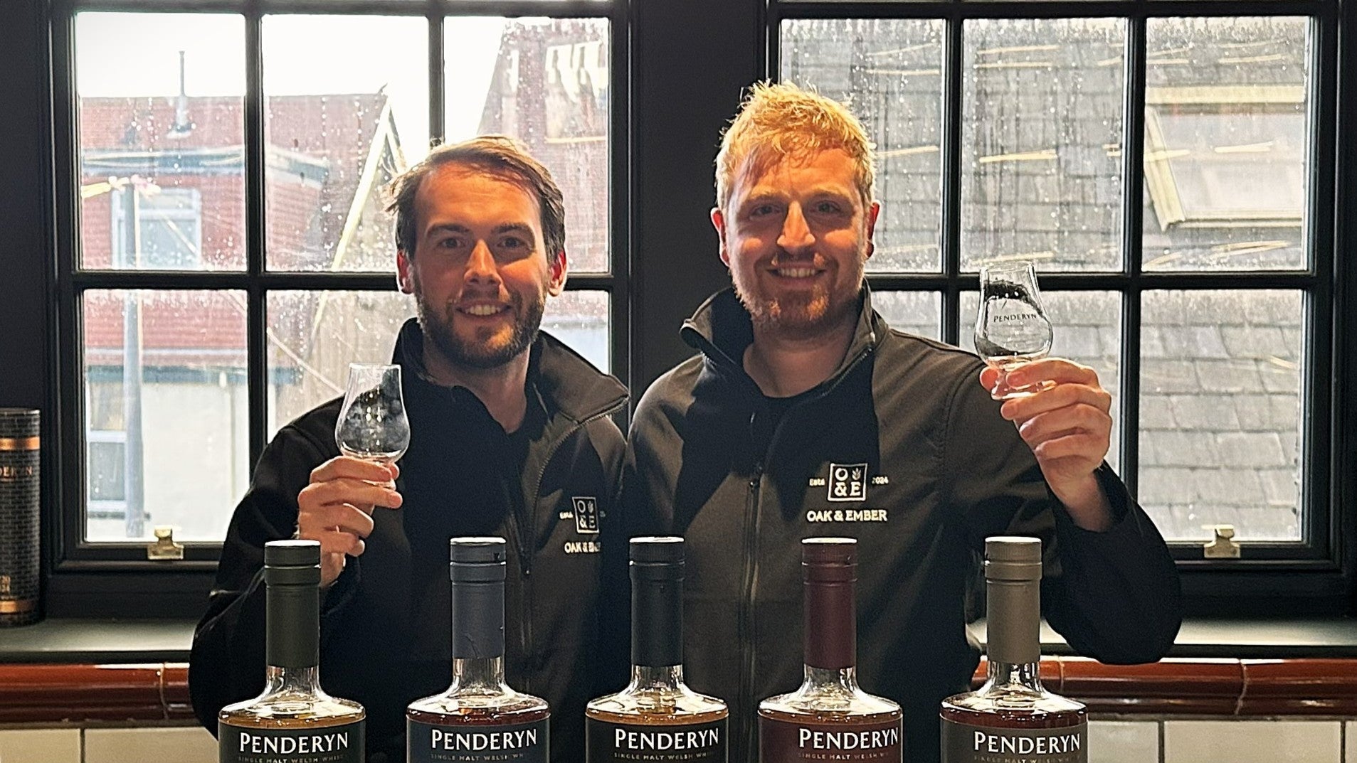 Two Oak & Ember team members raising whisky glasses during a Penderyn whisky tasting, standing behind bottles of single malt whisky.