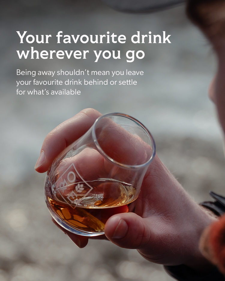 A person holds a small clear glass tumbler with amber liquid in it, with large white marketing text above reading 'Your favourite drink wherever you go' and additional copy about keeping your favourite drink when away.