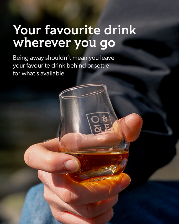 A hand holds a small clear glass tumbler with amber liquid in it outdoors, with overlaid marketing text about always having your favourite drink wherever you go. The glass has a visible square logo reading O & F.