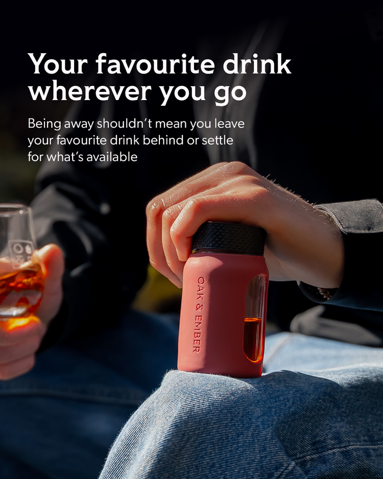 A person holds a small pink insulated bottle with a black lid while sitting outdoors in bright sunlight. The bottle has a clear side window showing amber liquid inside, and the brand name OAK & ENBER is printed vertically on the front.